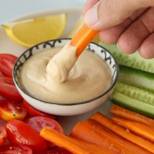 A hand dips a carrot stick into a bowl of creamy tahini dip, surrounded by sliced cherry tomatoes, carrots, cucumber, and a lemon wedge on a plate.