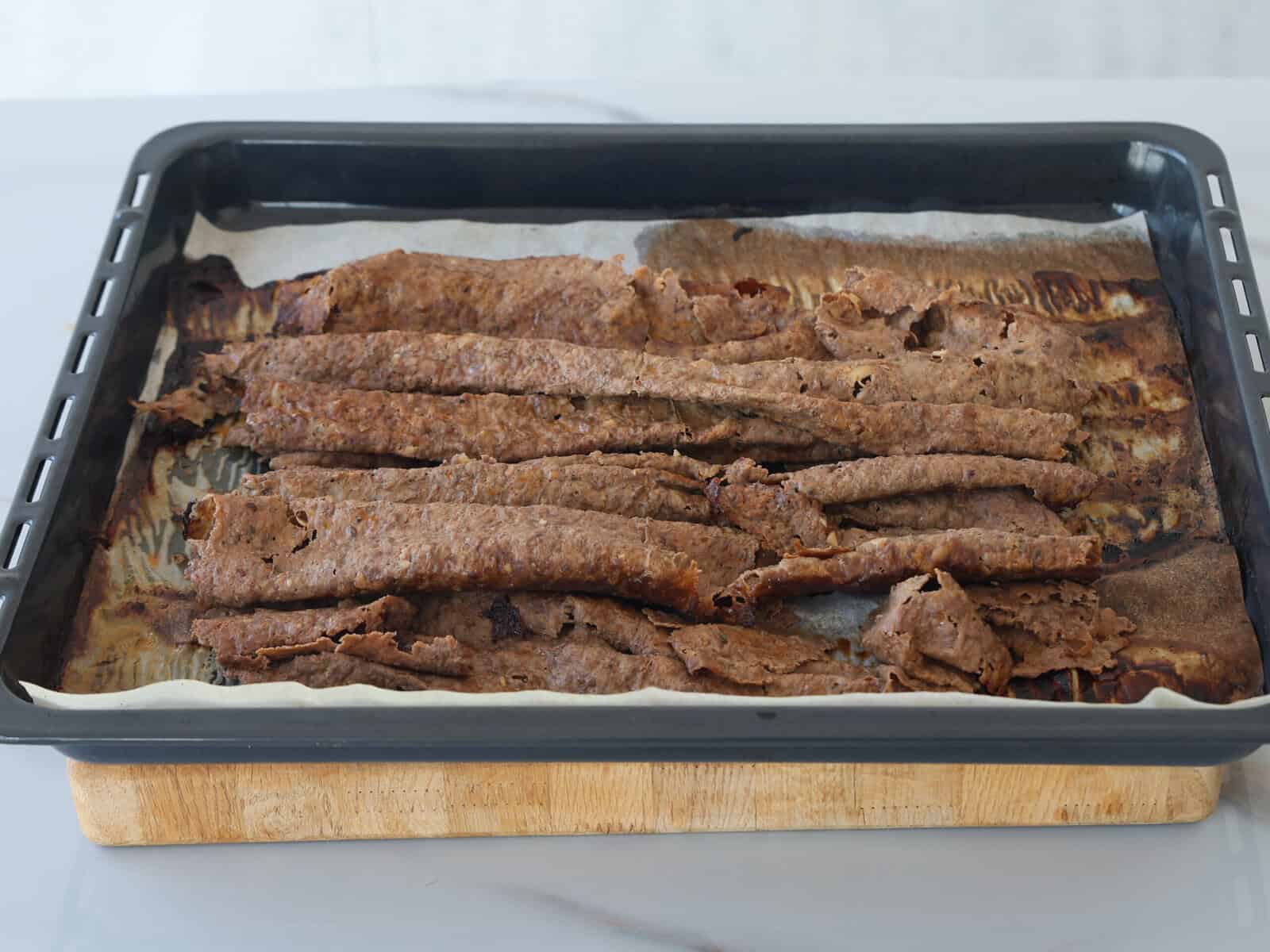 A baking tray lined with parchment paper holds several strips of cooked, unevenly shaped brown meat, resting on a wooden board on a light-colored surface.