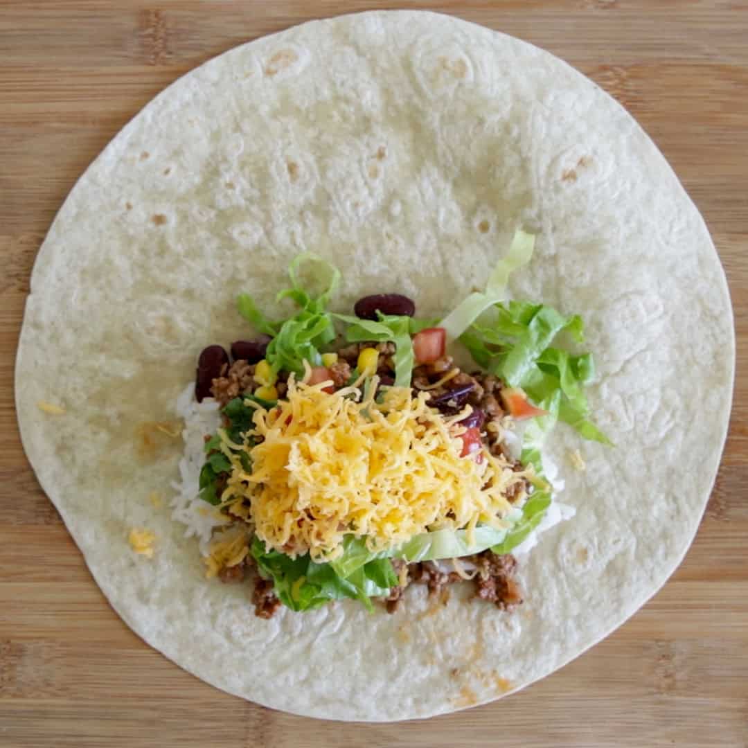 A flour tortilla on a wooden surface topped with rice, ground meat, kidney beans, chopped tomatoes, shredded lettuce, and grated cheese, ready to be wrapped as a burrito.