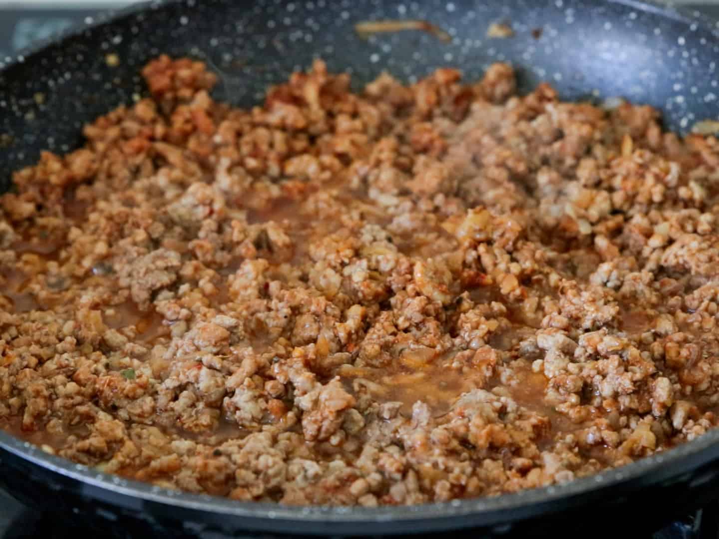 Ground meat is cooking in a skillet with visible juices and browned bits, creating a savory mixture often used for dishes like tacos or spaghetti sauce.
