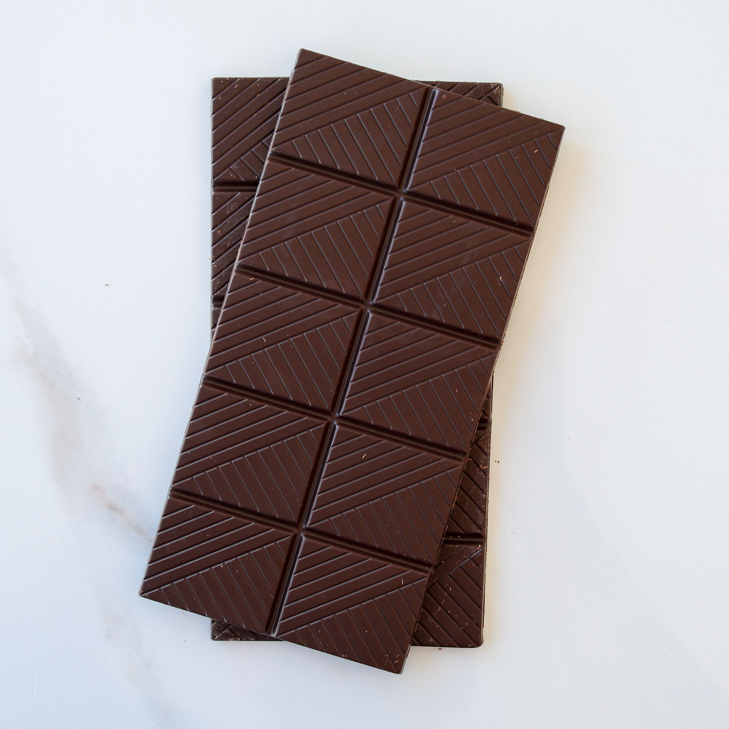 Two dark chocolate bars with a geometric pattern of diagonal lines, stacked slightly askew on a white surface.