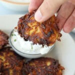 A hand dips a crispy, golden-brown potato fritter into a bowl of creamy white sauce on a plate with more fritters.