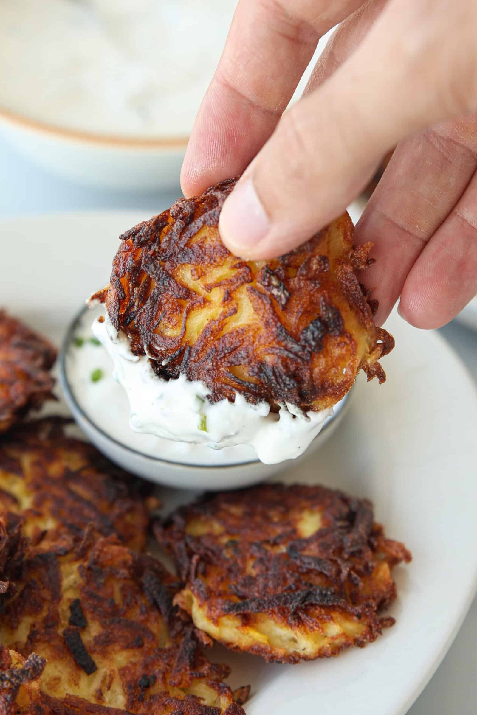 A hand dips a crispy potato fritter into a bowl of creamy white sauce. More fritters are stacked on a white plate nearby.