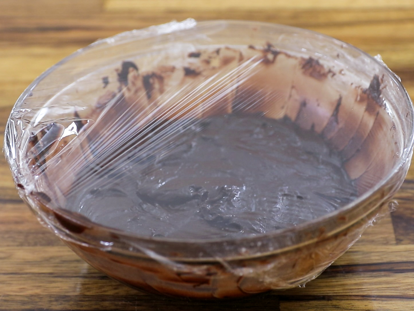 A glass bowl filled with chocolate batter covered in plastic wrap, resting on a wooden surface.