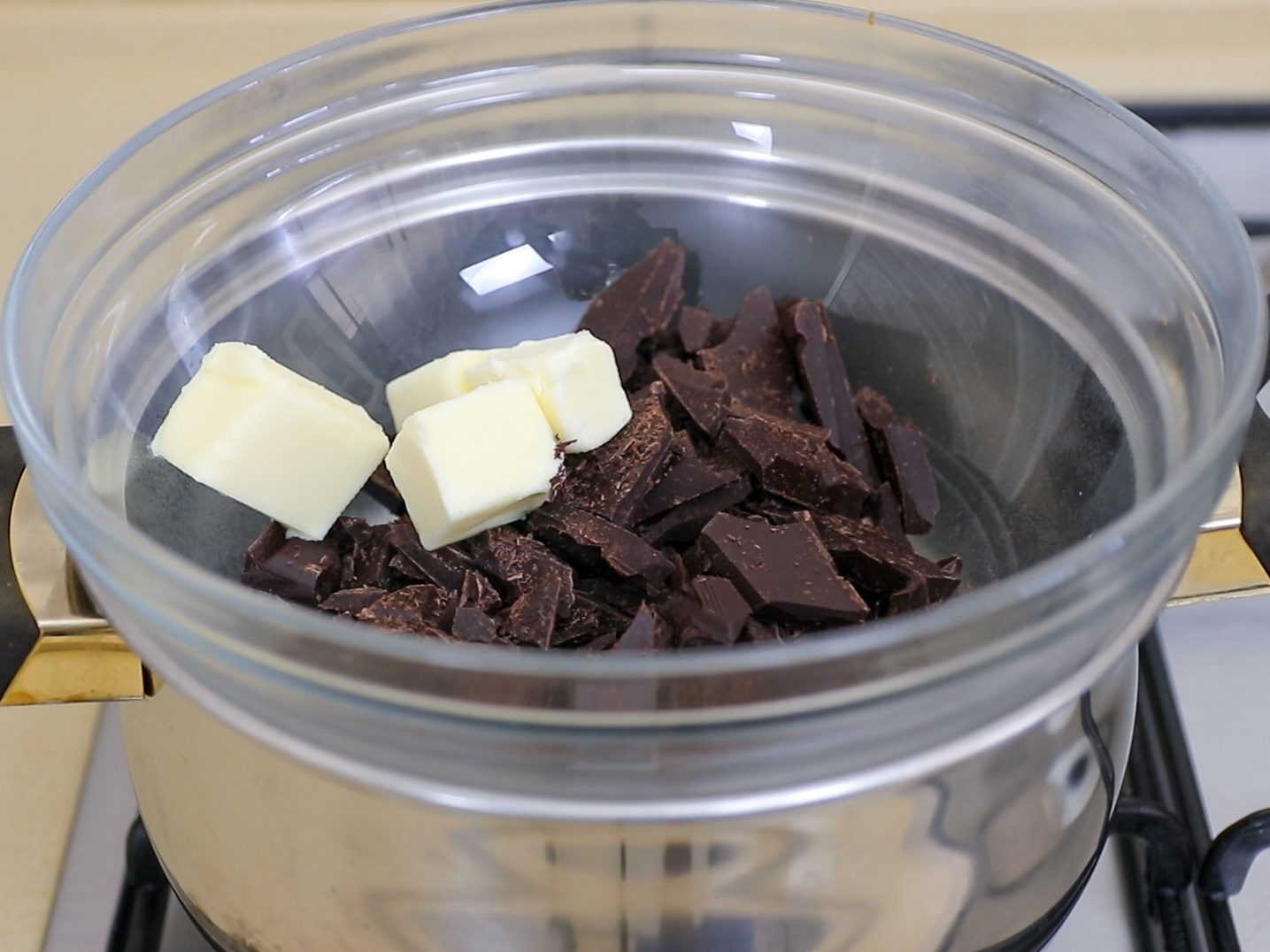 A glass bowl with chunks of dark chocolate and pieces of butter sits on top of a pot filled with hot water, forming a double boiler on a stove, ready for melting.