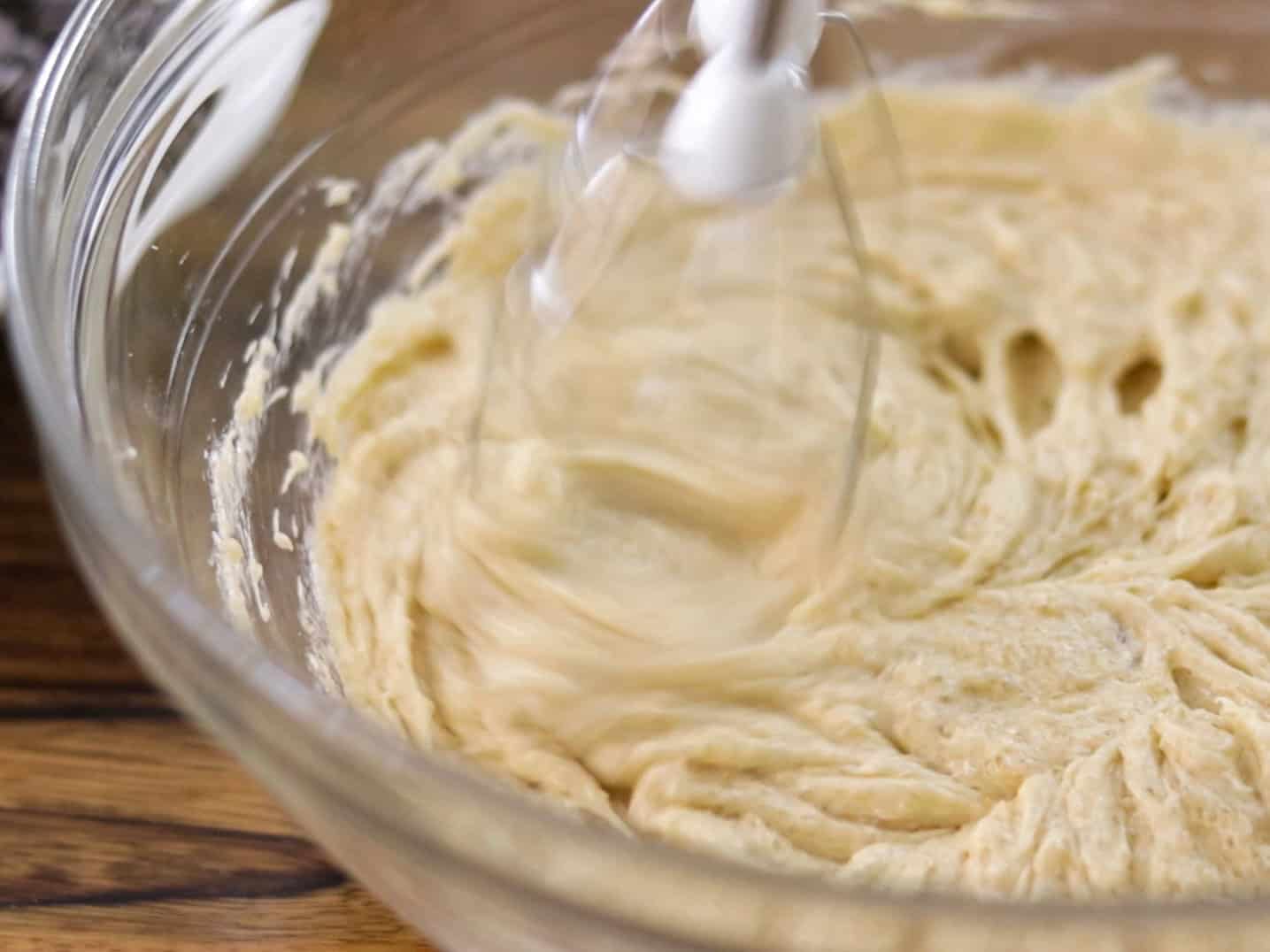 A close-up of an electric hand mixer blending creamy batter in a clear glass bowl on a wooden surface. The motion of the mixer blades creates swirling patterns in the batter.
