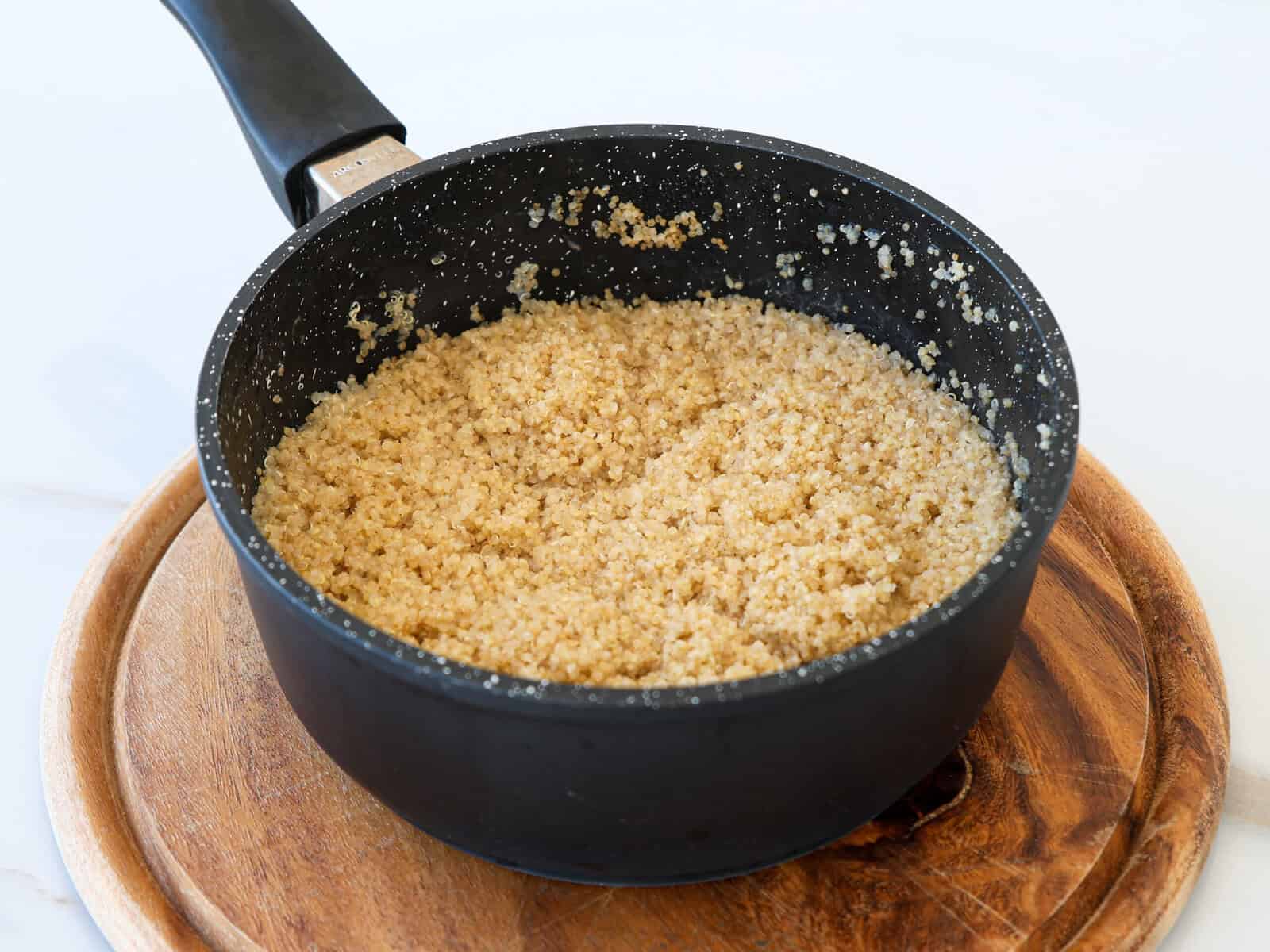A black saucepan filled with cooked quinoa sits on a round wooden cutting board against a white background.