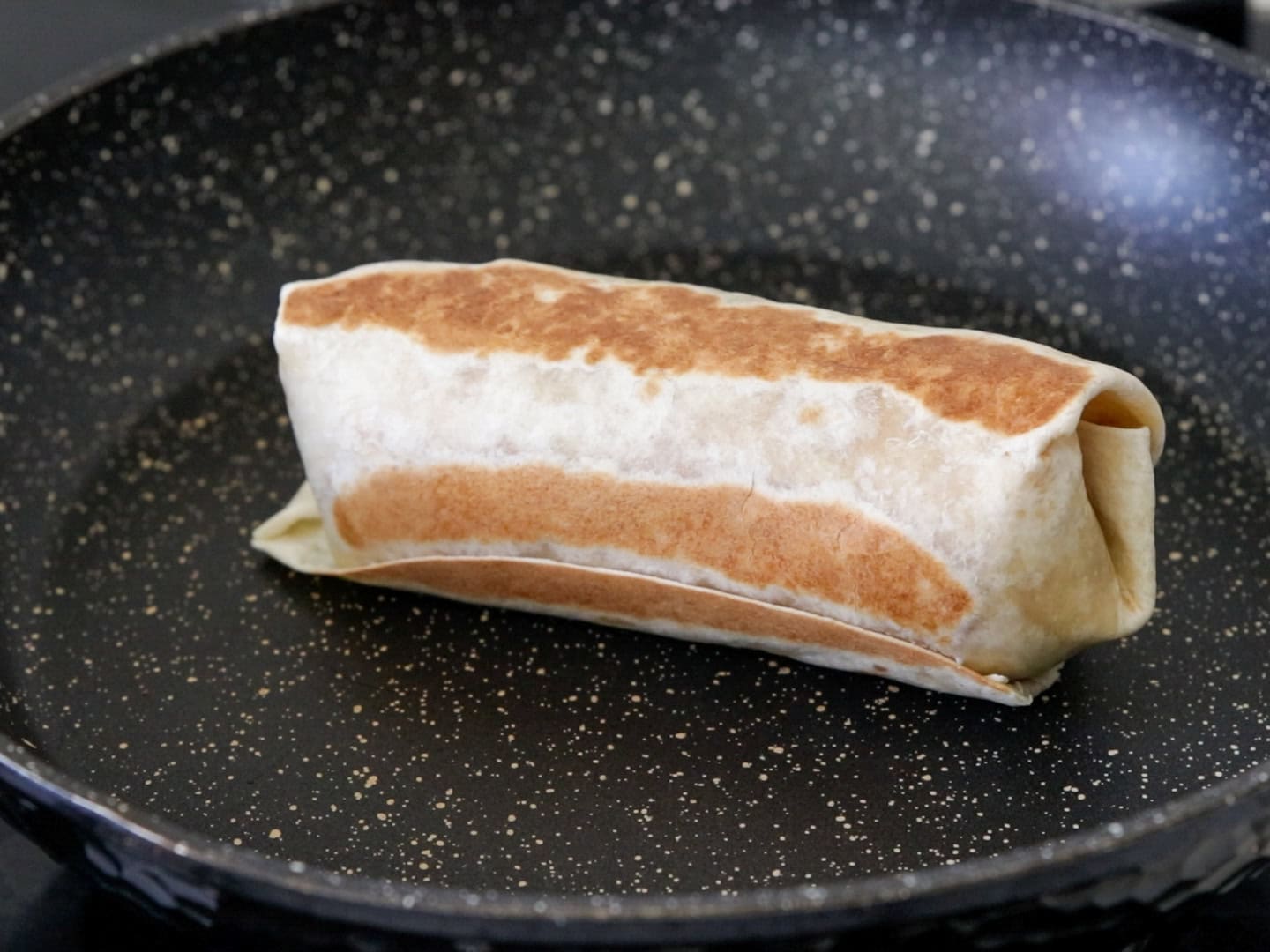 A browned burrito or wrap is being cooked in a nonstick frying pan with a speckled surface.