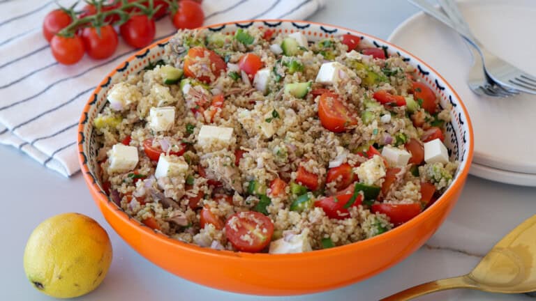 A large orange bowl filled with quinoa and tuna salad, featuring cherry tomatoes, cucumber, feta cheese cubes, herbs, and tuna. A lemon, tomatoes on the vine, plates, and utensils are nearby on a striped cloth.