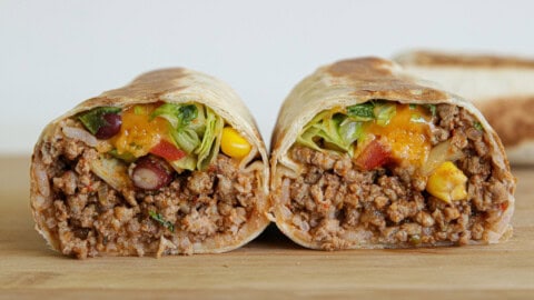 A close-up of a burrito cut in half, showing a filling of seasoned ground beef, rice, beans, corn, lettuce, diced tomatoes, and melted cheese, all wrapped in a flour tortilla on a wooden surface.