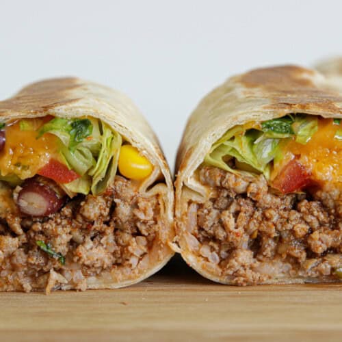 A close-up of a burrito cut in half, showing a filling of seasoned ground beef, rice, beans, corn, lettuce, diced tomatoes, and melted cheese, all wrapped in a flour tortilla on a wooden surface.