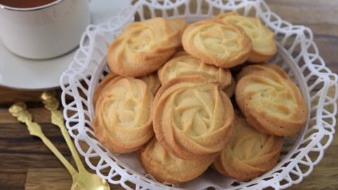 A white basket filled with round, golden Danish butter cookies sits on a wooden table beside a cup of tea and ornate gold spoons.