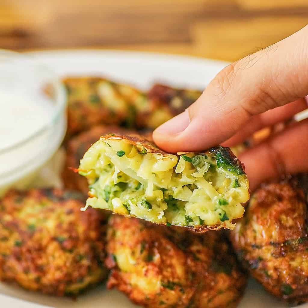 A hand holds a halved cabbage fritter, revealing its moist, green and yellow vegetable filling. Other fritters and a bowl of dipping sauce are visible on a white plate in the background.