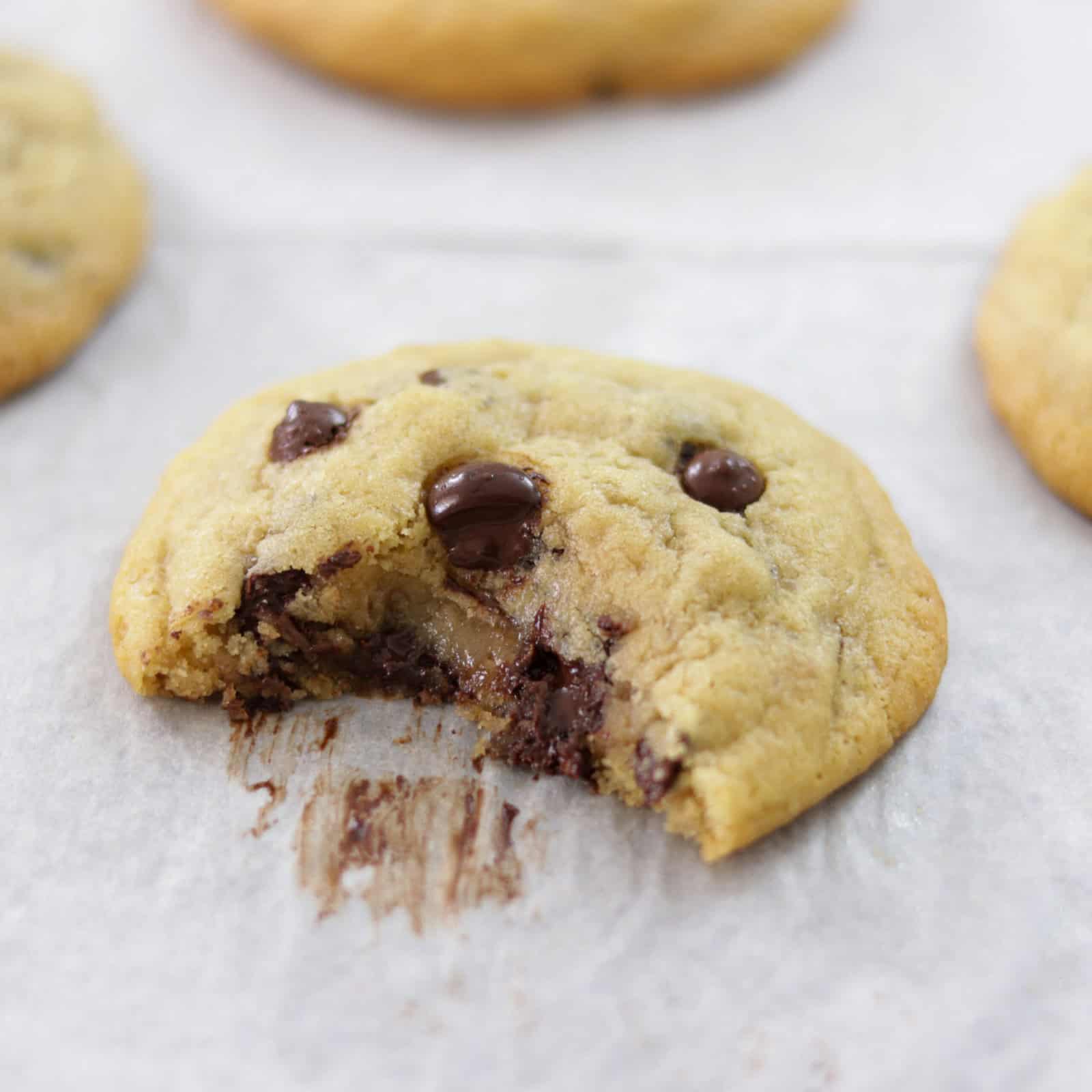 A close-up of a chocolate chip cookie with a bite taken out, resting on white parchment paper. Melted chocolate chips are visible, and other whole cookies are slightly blurred in the background.