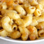 Close-up of creamy macaroni and cheese in a white bowl, with golden cheesy sauce coating the curved pasta and visible browned bits throughout.