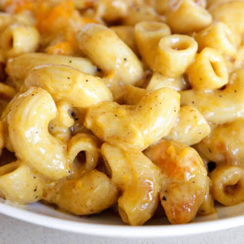 Close-up of creamy macaroni and cheese in a white bowl, with golden cheesy sauce coating the curved pasta and visible browned bits throughout.