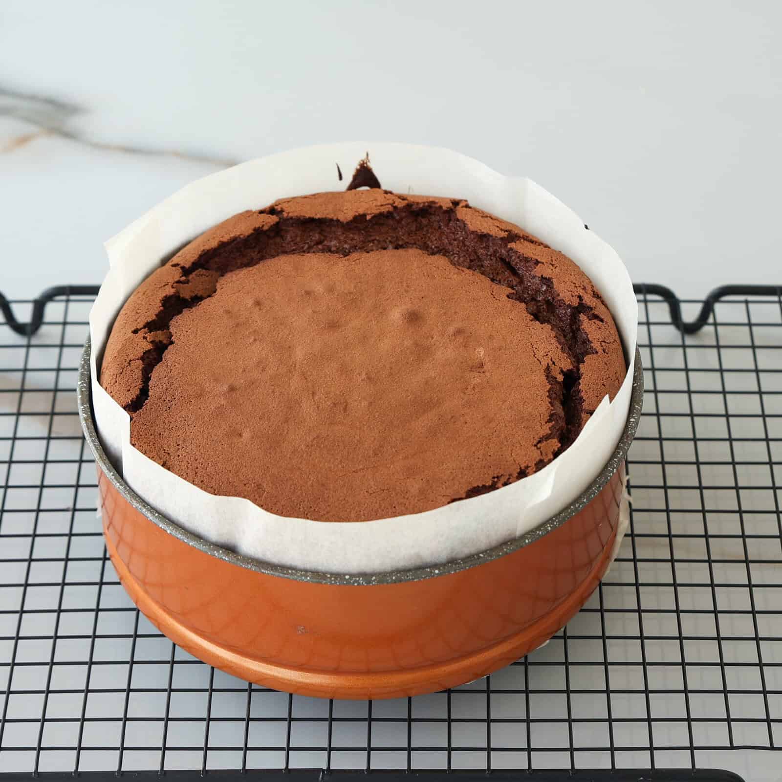 A freshly baked chocolate cake in a round pan lined with parchment paper rests on a black cooling rack. The cake has a cracked top and a rich brown color.