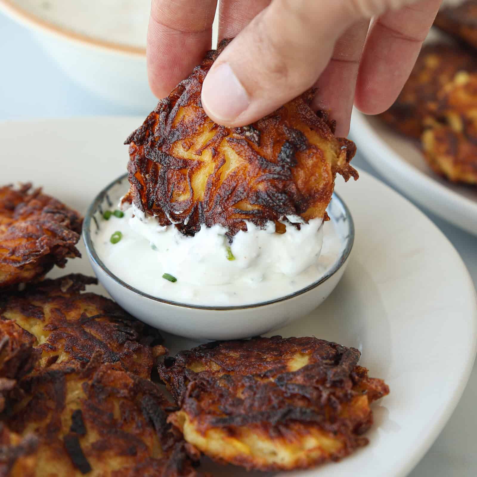 A hand dips a crispy, golden-brown potato fritter into a bowl of sour cream dip, herb-speckled sauce on a white plate with more fritters.