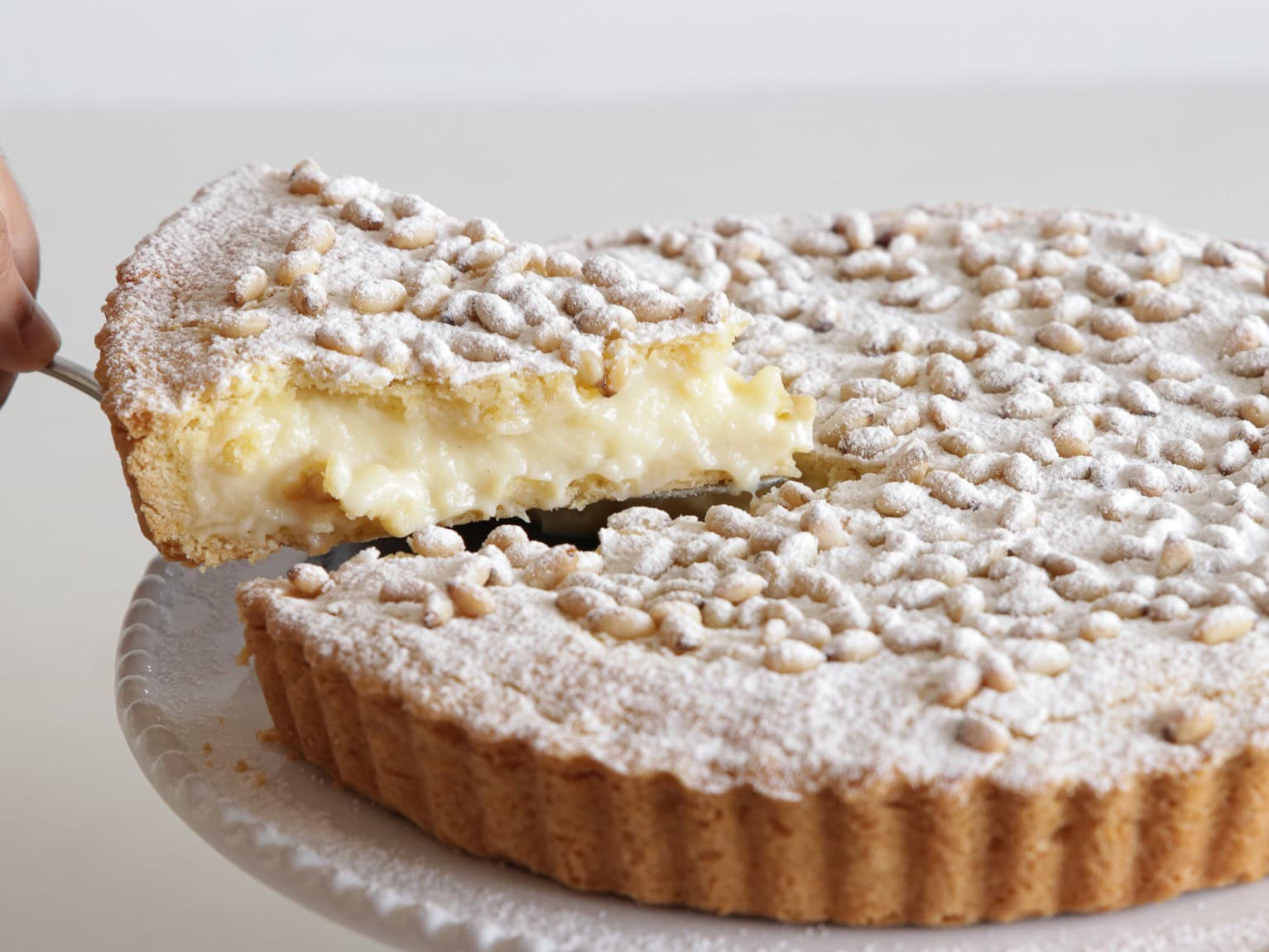 A hand lifts a slice of Italian pine nut tart (torta della nonna) from a whole tart. The tart has a golden crust, creamy filling, powdered sugar, and pine nuts on top, displayed on a white cake stand.