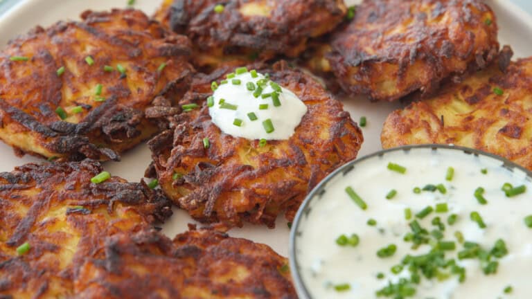 Crispy potato latkes topped with sour cream and chopped chives are arranged on a plate, with a bowl of creamy dip garnished with chives on the side.