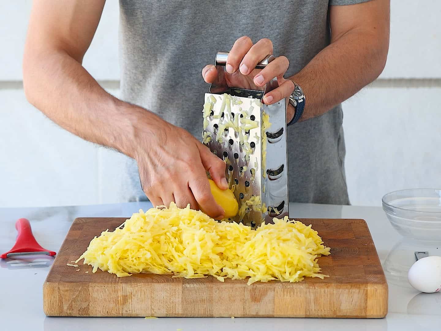 A person grates a potato on a box grater over a wooden cutting board, creating a pile of shredded potato. A peeler, a glass bowl, and two eggs are on the white countertop nearby.