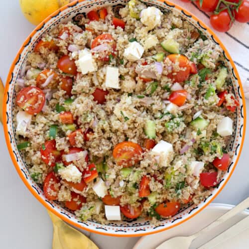 A colorful bowl of bulgur salad with cherry tomatoes, cucumber, feta cheese cubes, and red onions sits near a striped towel, fresh tomatoes on the vine, a lemon, a golden spoon, and two forks on a white plate.