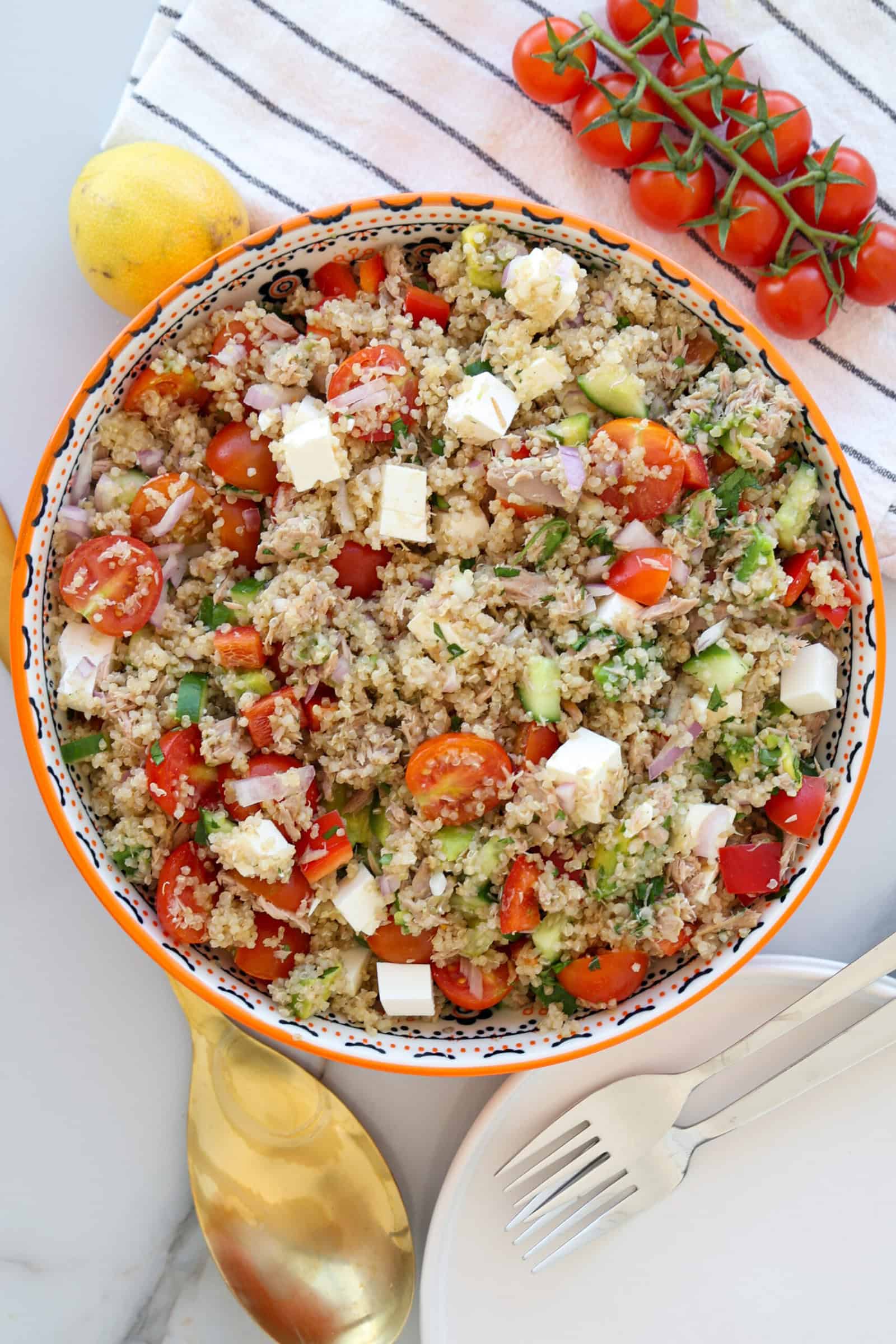 A colorful bowl of quinoa salad with tuna, cherry tomatoes, cucumber, feta cheese cubes, and red onions sits near a striped towel, fresh tomatoes on the vine, a lemon, a golden spoon, and two forks on a white plate.