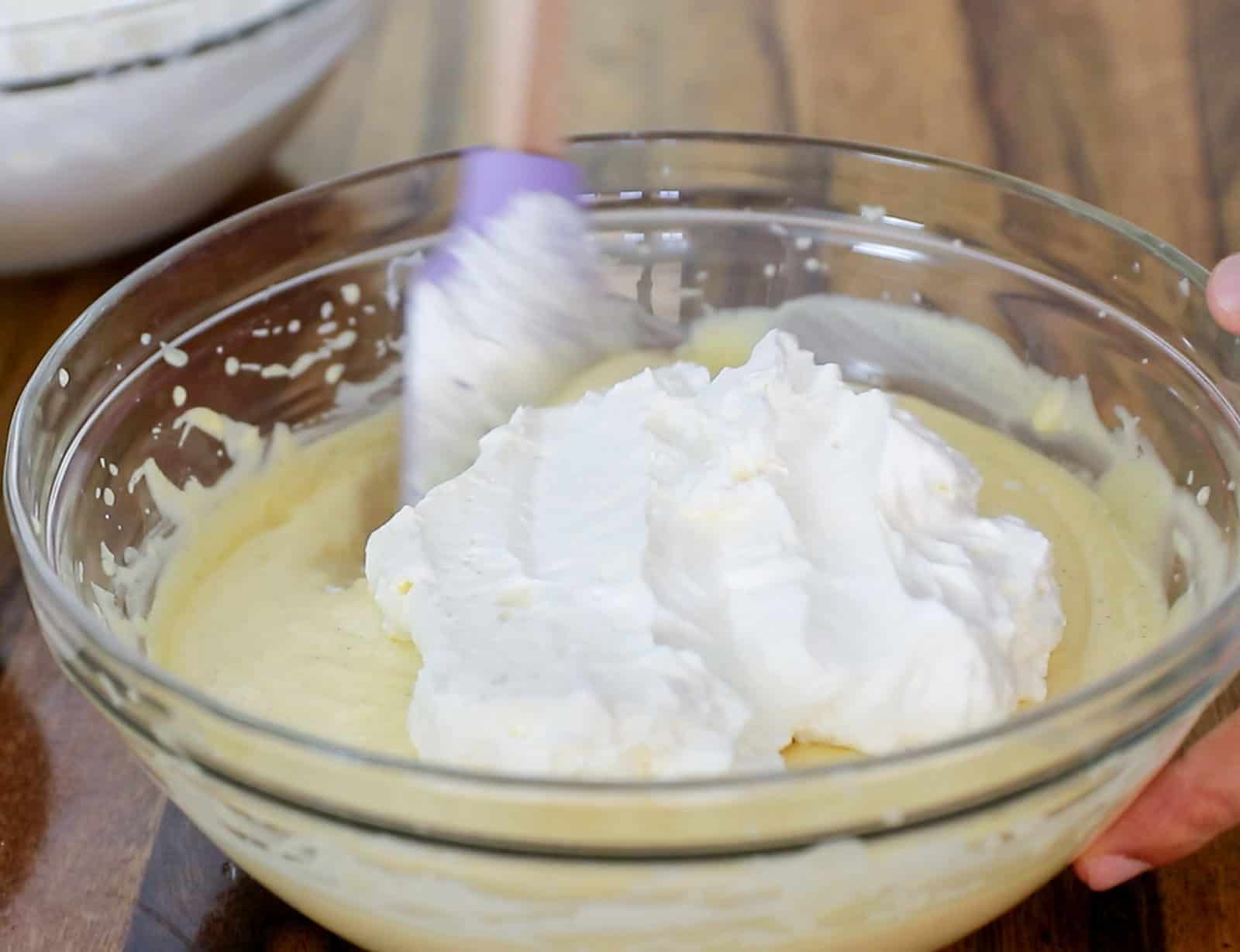 A close-up of a person using a spatula to fold whipped cream into a yellow batter in a glass mixing bowl on a wooden surface. Another bowl is visible in the background.