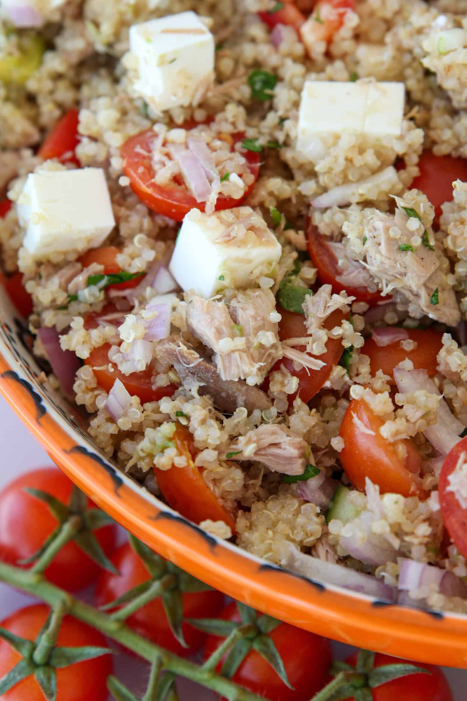 A close-up of a quinoa salad with cherry tomatoes, cubed feta cheese, red onion, herbs, and chunks of tuna in an orange bowl, with a vine of fresh tomatoes beside it.
