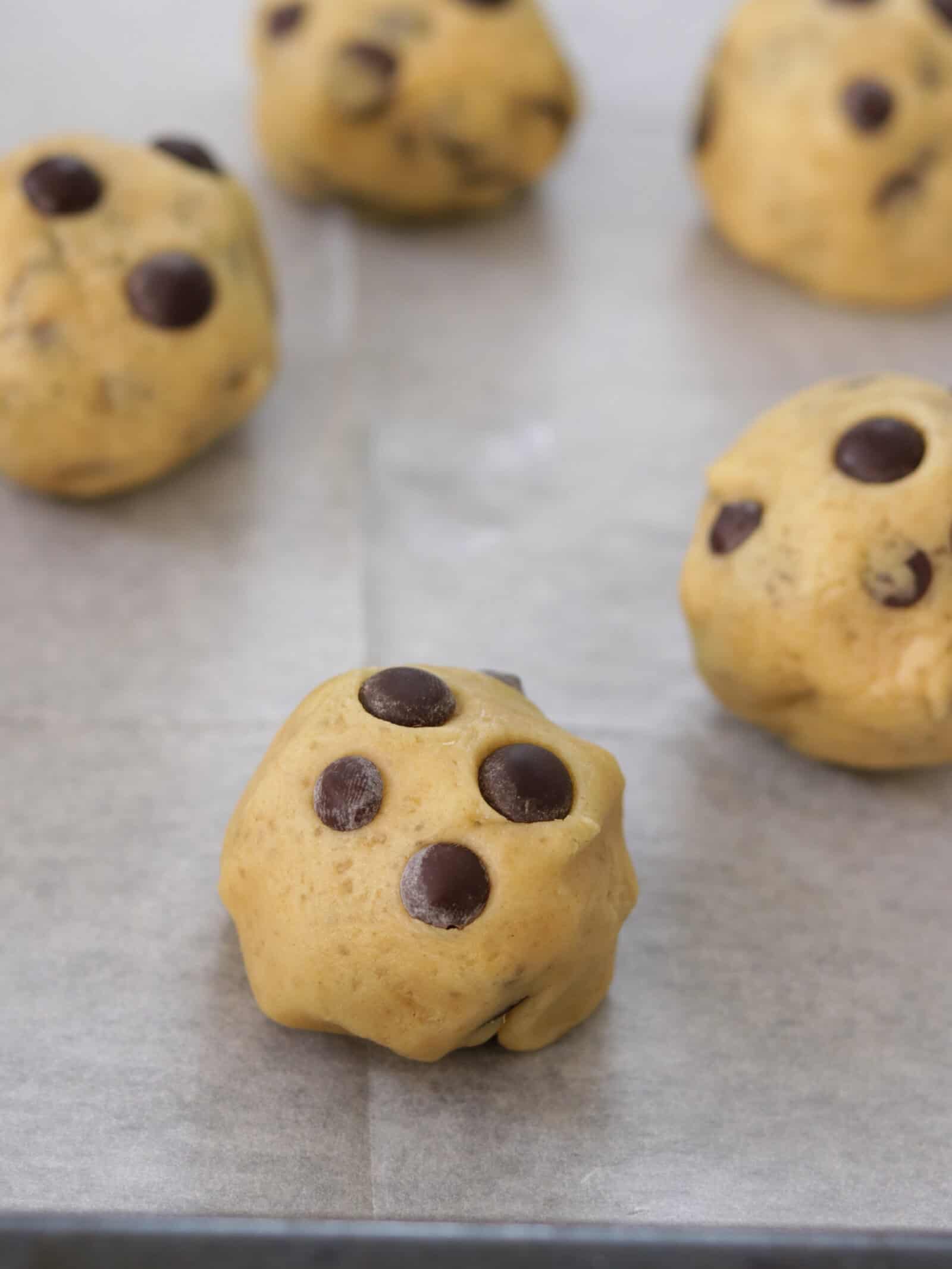 Close-up of several balls of chocolate chip cookie dough on a parchment-lined baking sheet, with chocolate chips visible on the dough surface.