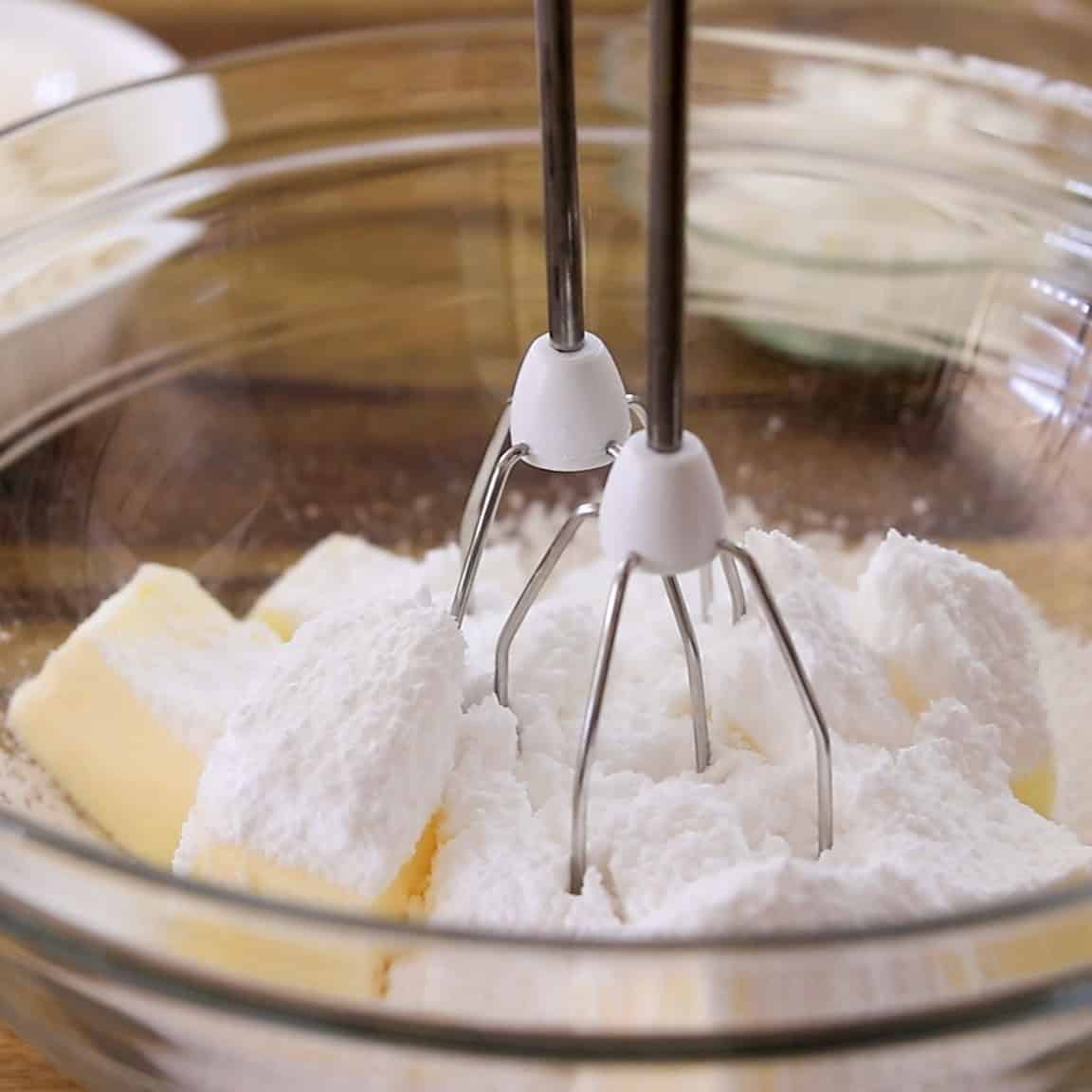 Two metal beaters of a hand mixer are placed in a glass bowl containing sticks of butter and a mound of powdered sugar, ready to be mixed.