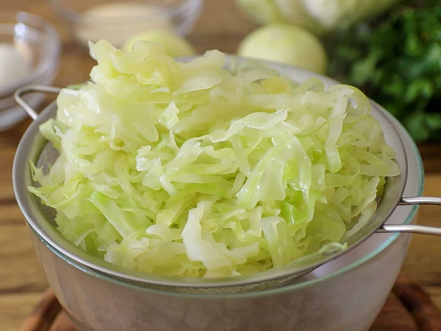 Shredded cabbage sits in a metal strainer placed over a glass bowl. The background shows blurred ingredients, including greens and white vegetables, on a wooden surface.