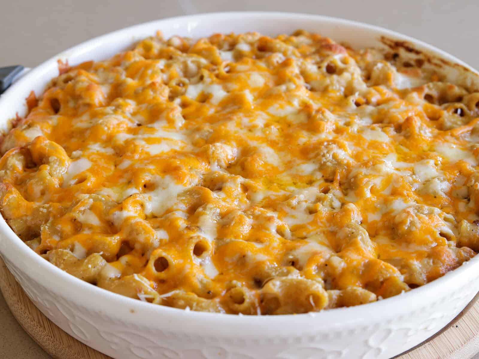 A baked macaroni and cheese casserole in a round white dish, topped with melted cheddar and mozzarella cheese, sitting on a wooden trivet on a countertop.