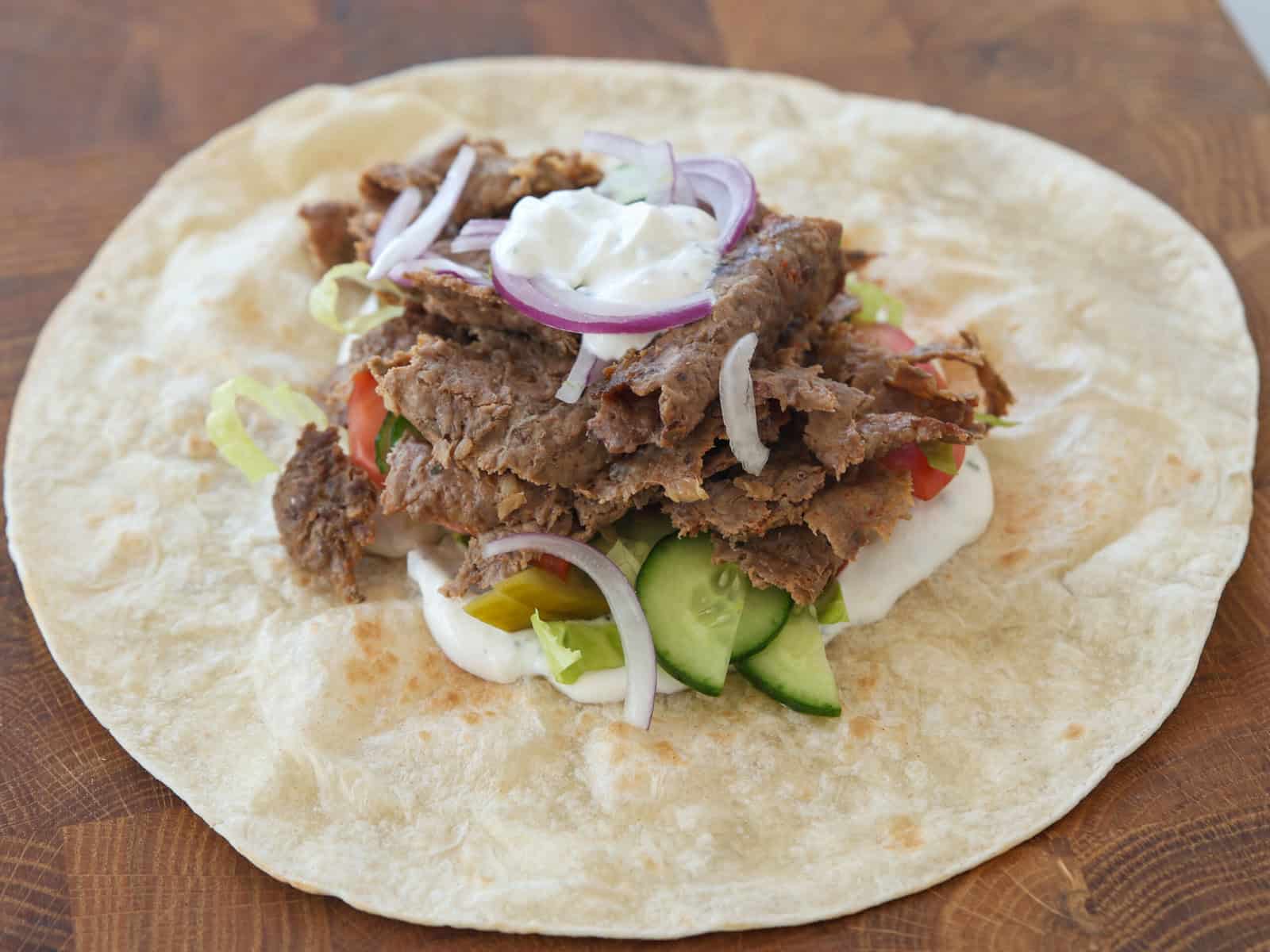 An open flatbread wrap topped with sliced meat, lettuce, cucumbers, tomatoes, red onion, and a dollop of white sauce, all placed on a wooden surface.