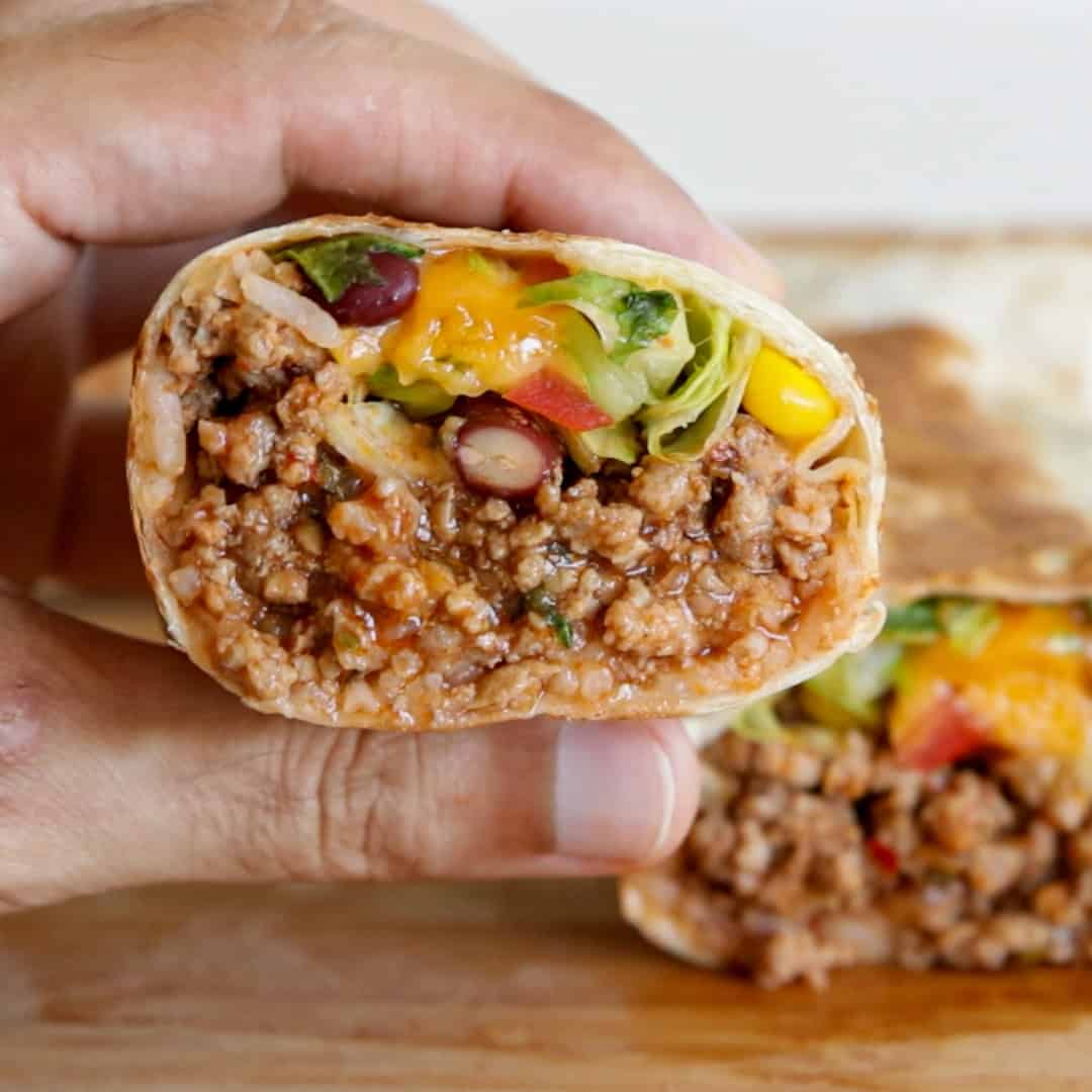 A hand holds a ground beef burrito cut in half, showing a filling of ground meat, rice, beans, lettuce, tomato, and melted cheese. Another half sits on a wooden surface in the background.