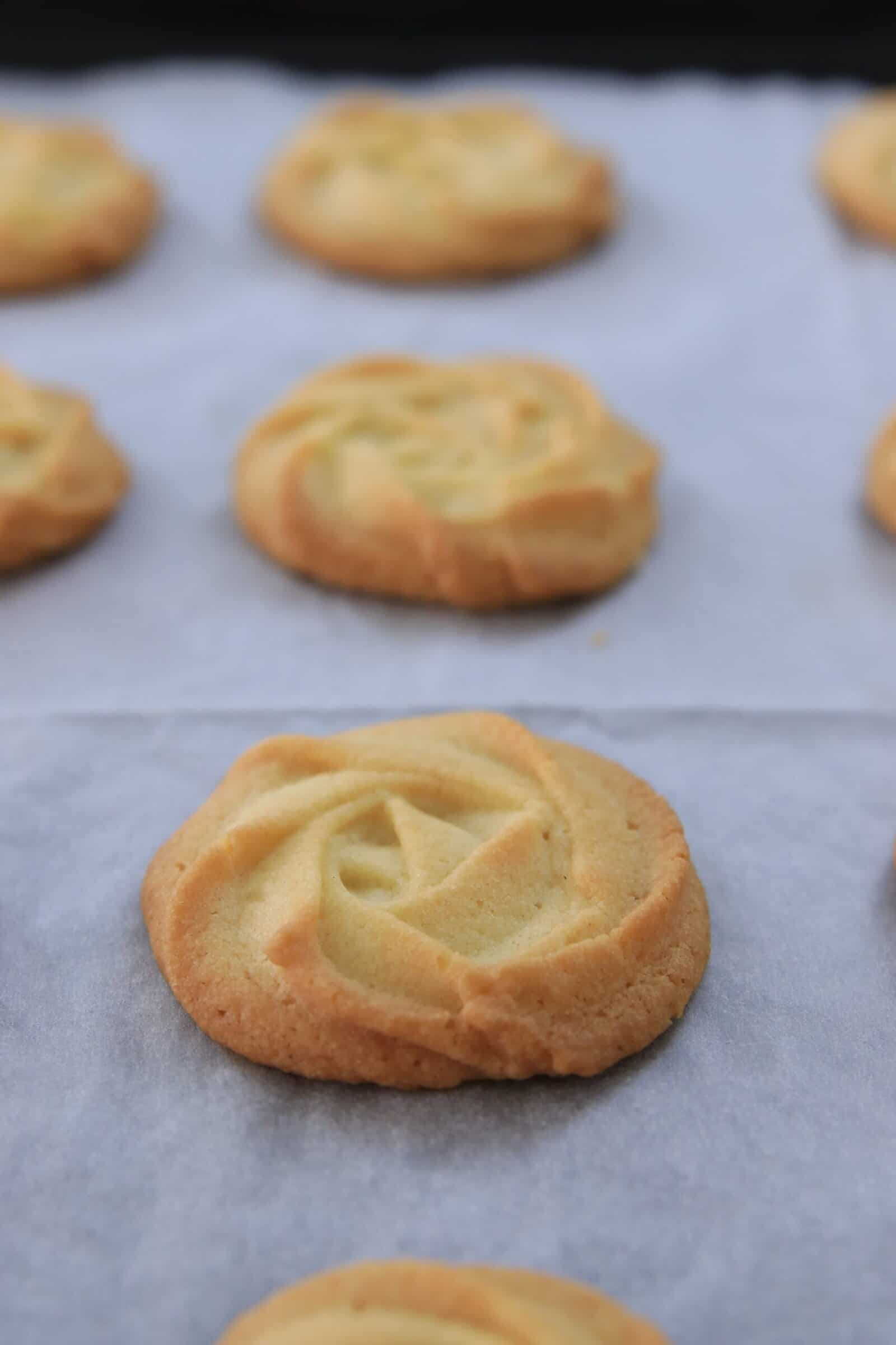 Close-up of several golden, round butter cookies with a swirled pattern, arranged on a sheet of white parchment paper. The cookies look freshly baked and slightly browned at the edges.