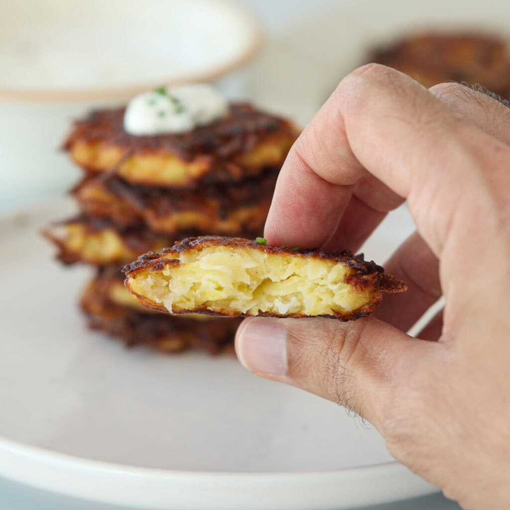 A hand holding a halved potato latke showing its crispy, golden-brown exterior and soft, shredded potato interior. Blurred latkes and a dollop of sour cream with chives are visible in the background.
