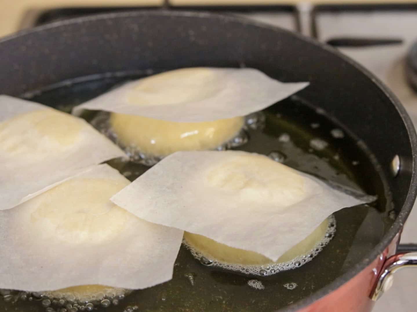 Four pieces of sufganiyot dough covered with parchment paper frying in hot oil in a deep pan on a stovetop.