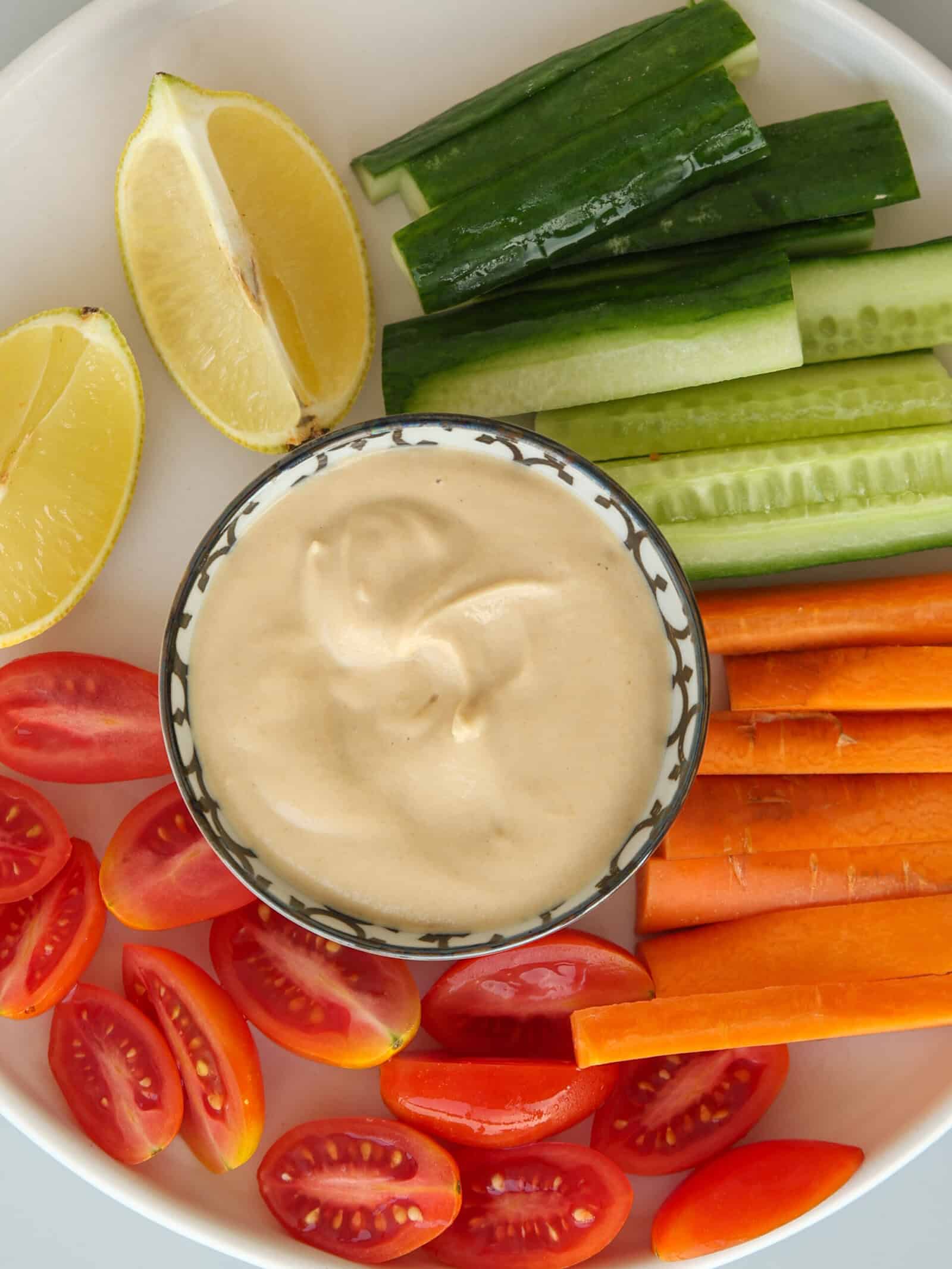 A plate with sliced cucumbers, carrots, grape tomatoes, lemon wedges, and a bowl of creamy lemon tahini sauce in the center.