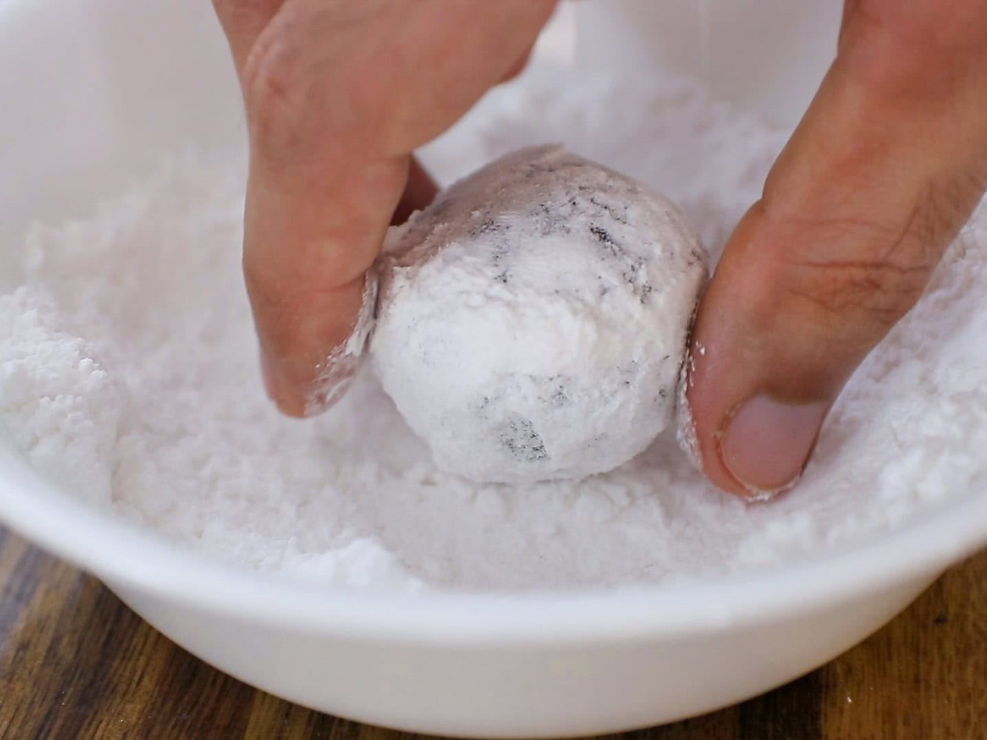 A hand rolls a round, chocolate cookie dough ball in a bowl filled with powdered sugar.