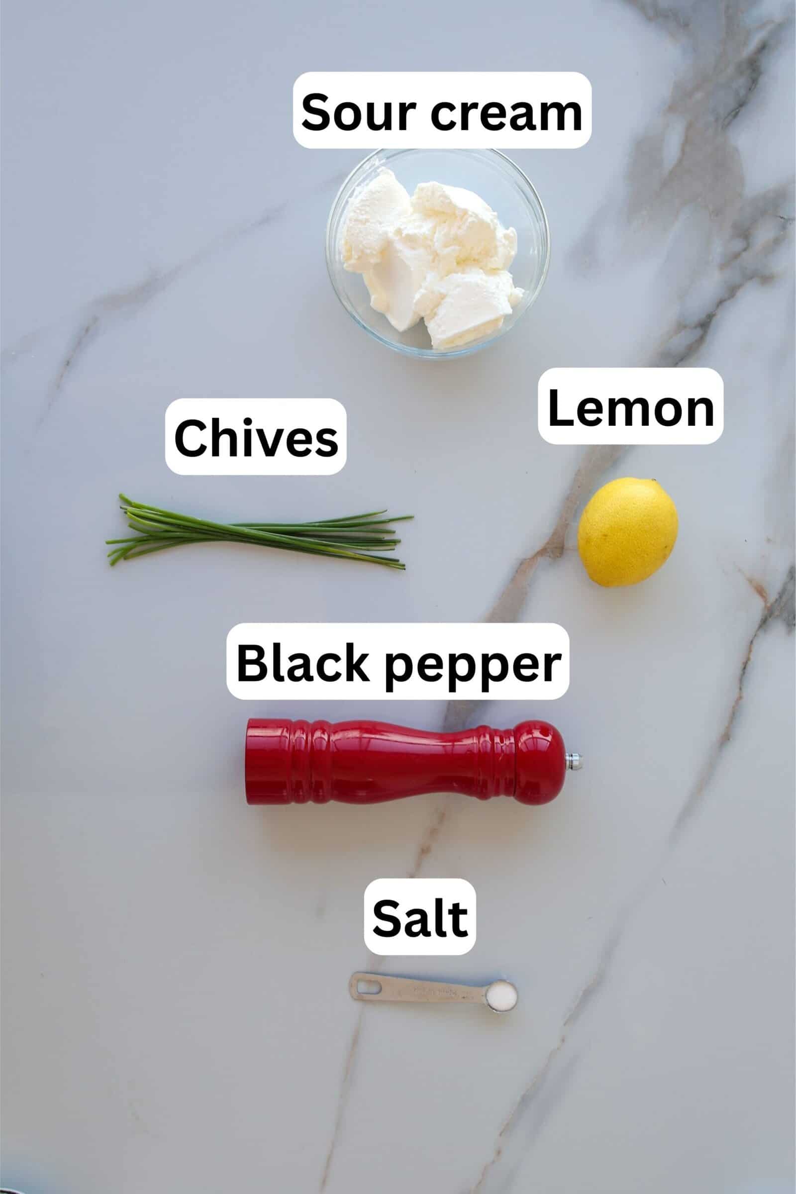 Ingredients on a white surface: a bowl of sour cream, whole lemon, bunch of chives, red pepper grinder, measuring spoon with salt. Each item is labeled with text describing its name.