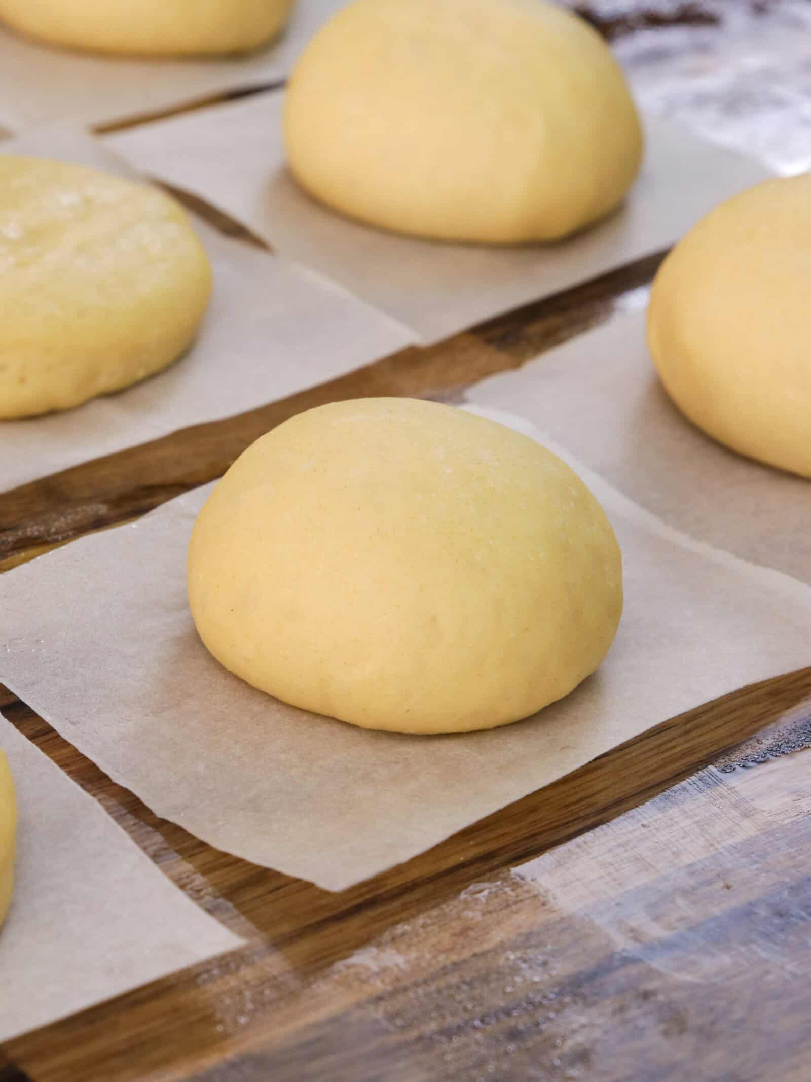 Balls of yellow dough rest on individual squares of parchment paper, spaced apart on a wooden surface, ready for baking or further preparation.