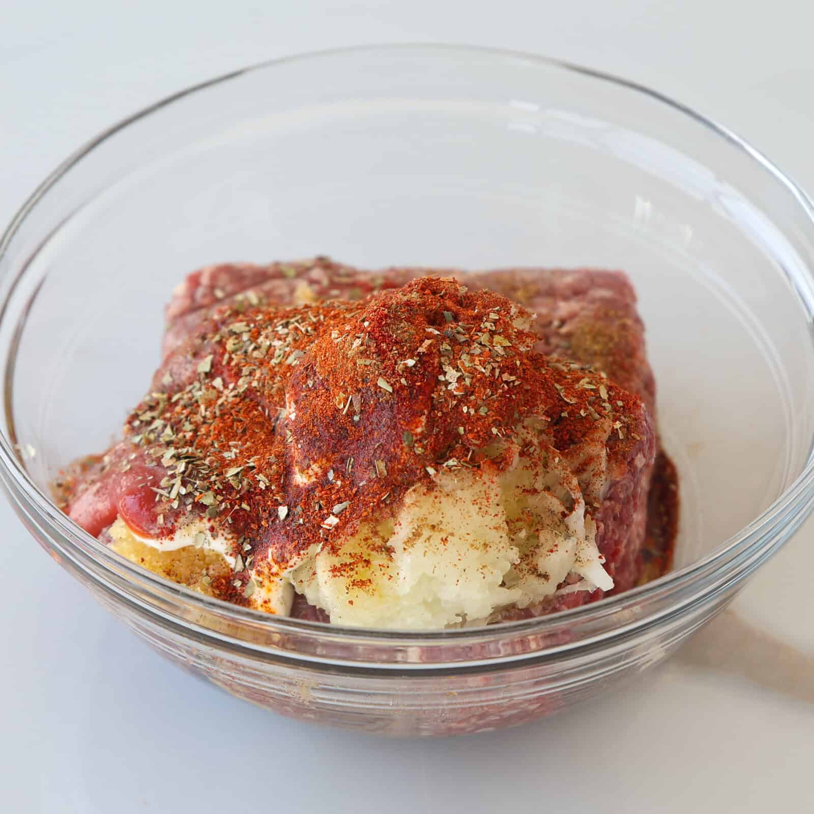 A glass bowl containing raw ground beef topped with shredded onions, paprika, dried herbs, and spices, all ready to be mixed together.
