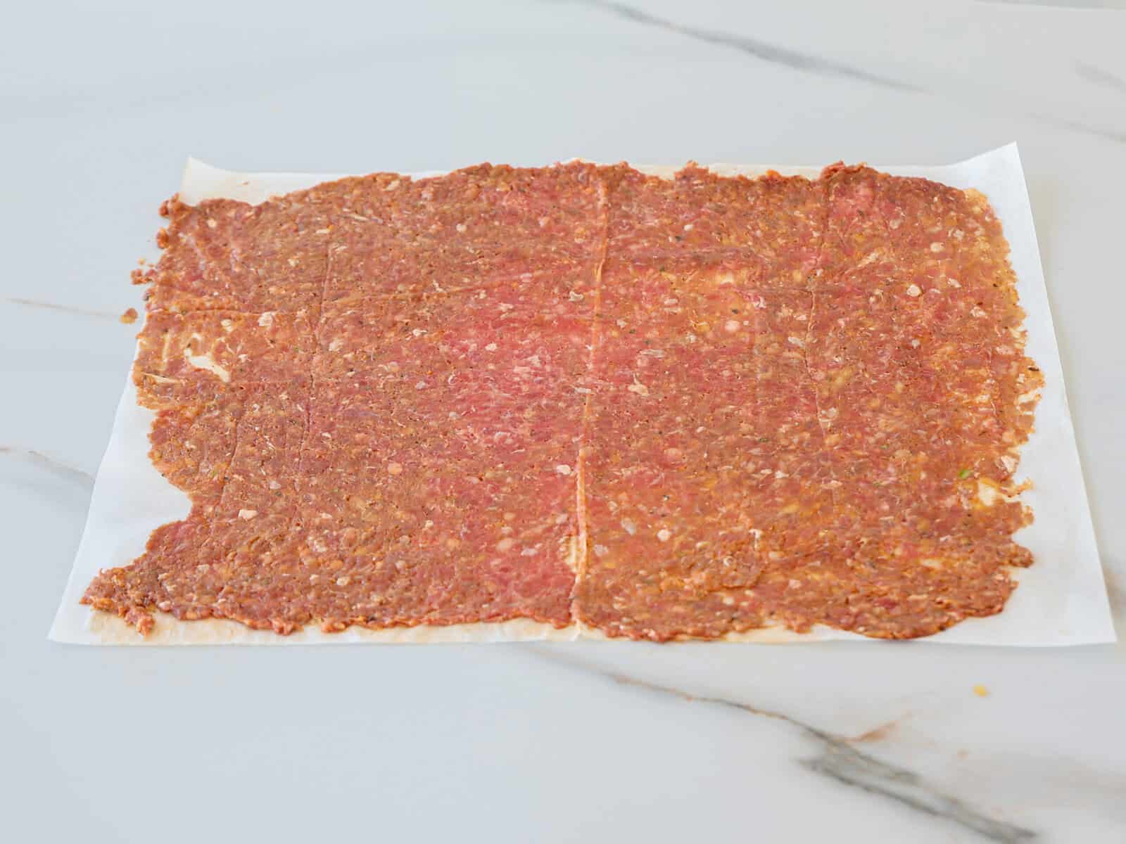 A sheet of ground meat mixture is spread thinly on parchment paper, resting on a white marble surface. The meat appears seasoned and is shaped into a rough rectangle with uneven edges.