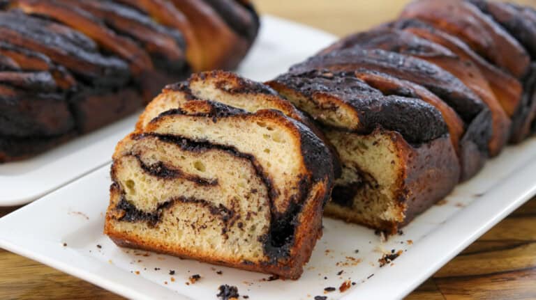 A close-up of sliced chocolate babka on a white rectangular plate, showing swirls of chocolate filling in the soft, golden bread. Another loaf is visible in the background.