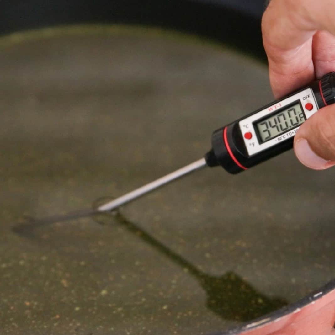 A hand holds a digital thermometer, measuring the temperature of a liquid in a pan. The display reads 34.0°C.