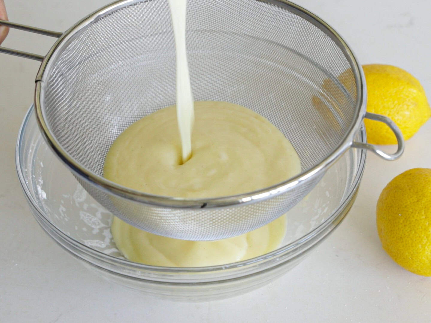 A creamy mixture is being poured through a fine mesh strainer into a glass bowl, with two whole lemons placed nearby on a white surface.