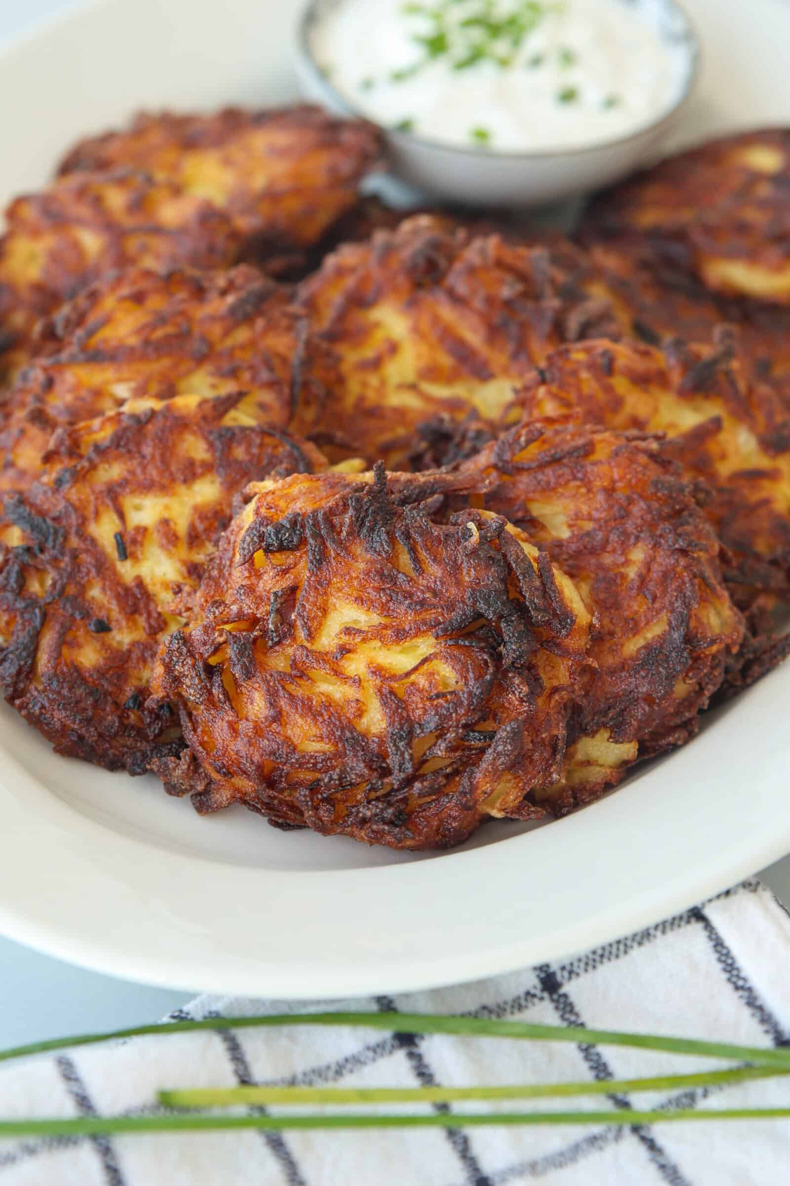A plate of crispy, golden-brown potato latkes with a small bowl of creamy sour cream dip garnished with chopped chives in the background. A striped towel and chives are visible in the foreground.