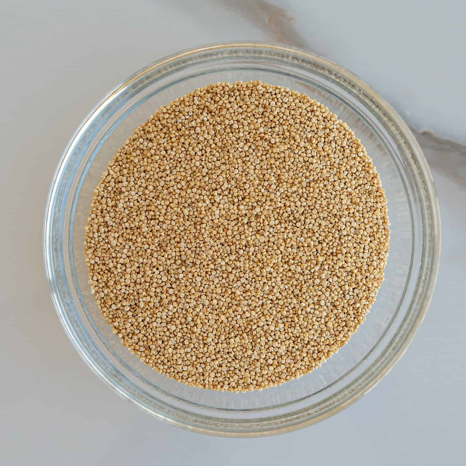 A clear glass bowl filled with raw, uncooked quinoa grains, placed on a white marble surface.