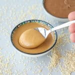 A spoon scoops creamy tahini from a small bowl, surrounded by scattered sesame seeds on a light surface. Another bowl with tahini is blurred in the background.